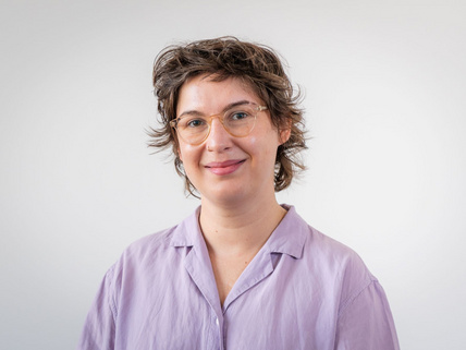 Portraitaufnahme von Charlotte Kunath. Sie steht vor einer grau-wei&szlig;en Wand und hat eine lilane Bluse an. Sie hat braune, leicht gewellte kurze Haare und tr&auml;gt eine Brille.