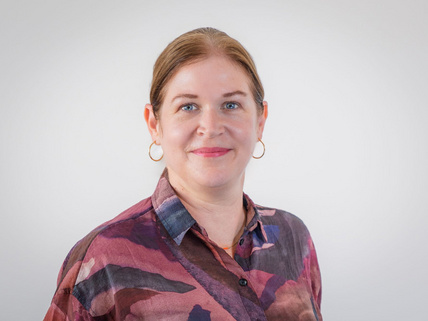 Portraitaufnahme von Sarah Schwarze: Sie steht vor einer wei&szlig;-grauen Wand, hat hellbraune Haare und tr&auml;gt einen Pferdeschwanz. Sie hat eine pink, lila gemusterte Bluse an, tr&auml;gt Ohrringe und rosafarbenen Lippenstift