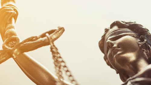 Nahaufnahme einer Statue der Justitia. Die Figur h&auml;lt eine Waage in der Hand, tr&auml;gt eine Augenbinde und ist in warmes Licht getaucht. Die Aufnahme ist leicht von unten fotografiert, sodass die Skulptur imposant wirkt.